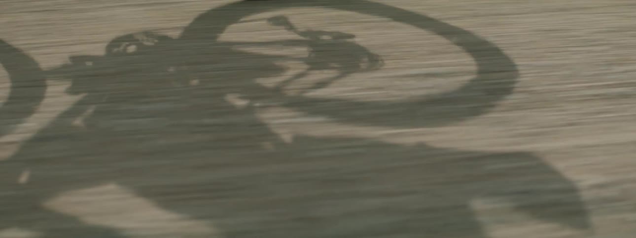 A person rides a bicycle down a gravel road with shadow cast