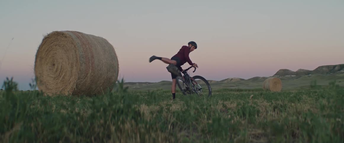 A man cycles across a field and gets off his bike next to a hay bale