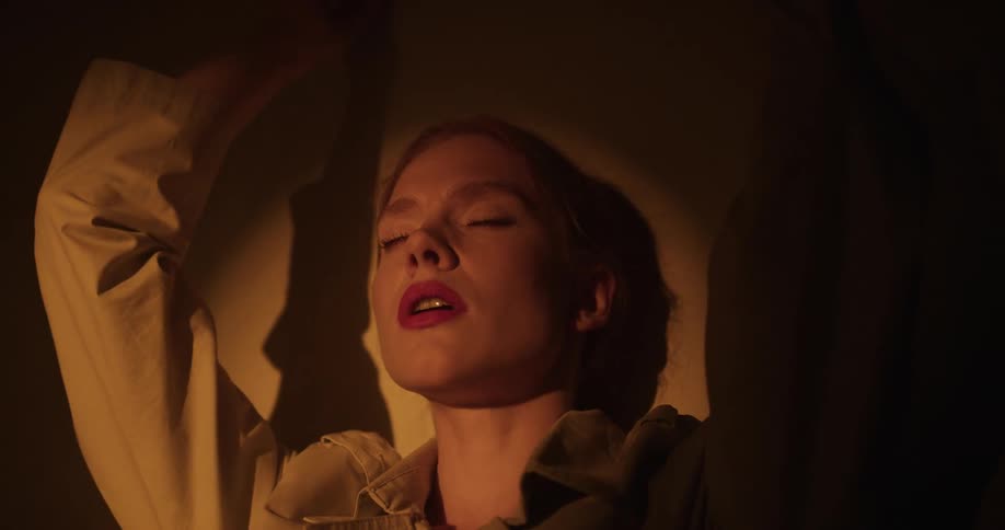 Female model with ponytail and brown overcoat closes eyes and spreads her arms in spotlight