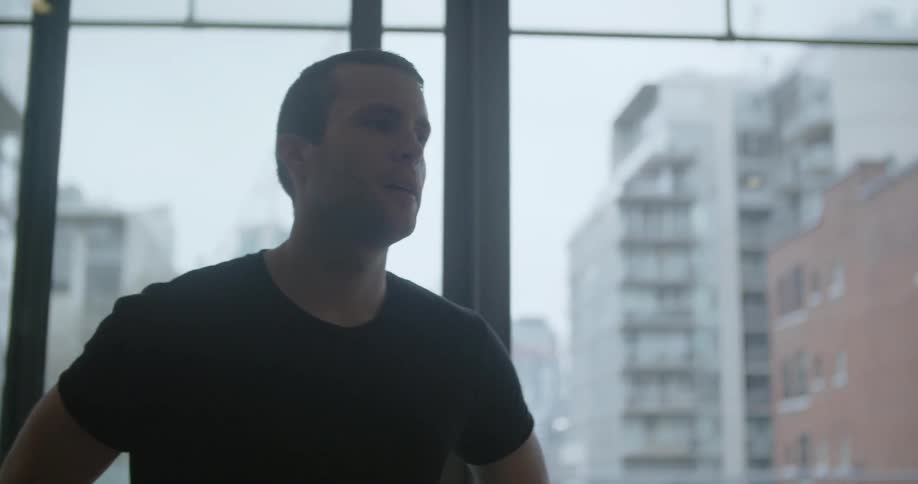 A man looking at the room around him as he stands next to windows that overlook tall buildings