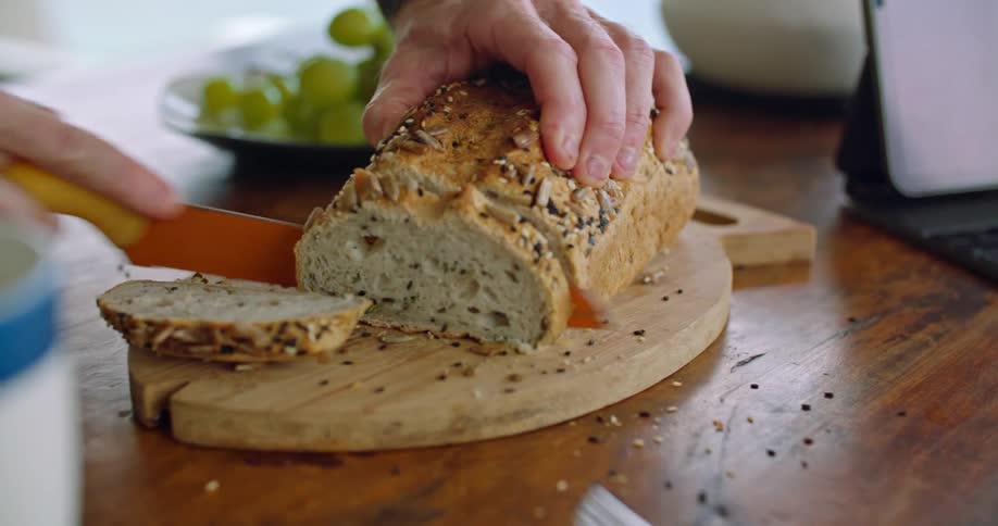 Man with watch cuts wholemeal bread with knife next to tablet and grapes on plate