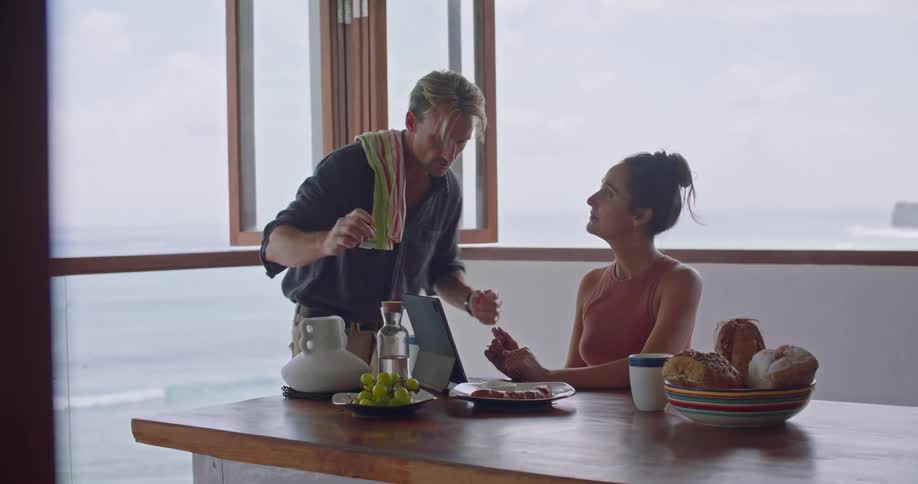 A woman sits at a table and types on a laptop and a man brings her a plate of food and bends down and they look at the laptop together
