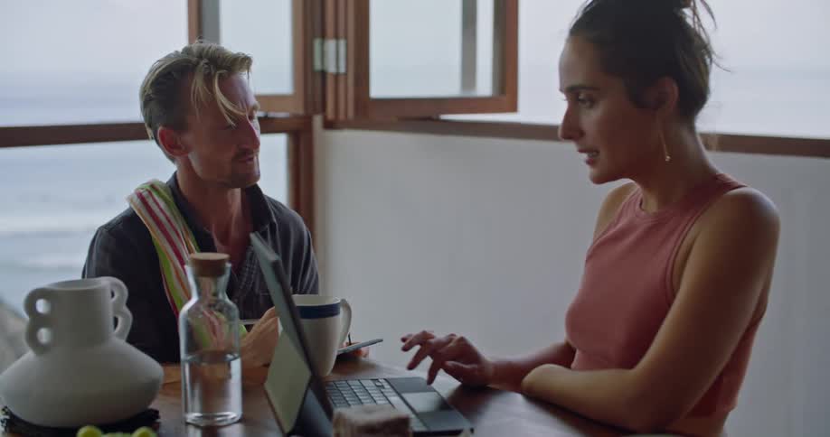 A man sits at a table and talks with a woman while she looks at a laptop and agrees with him