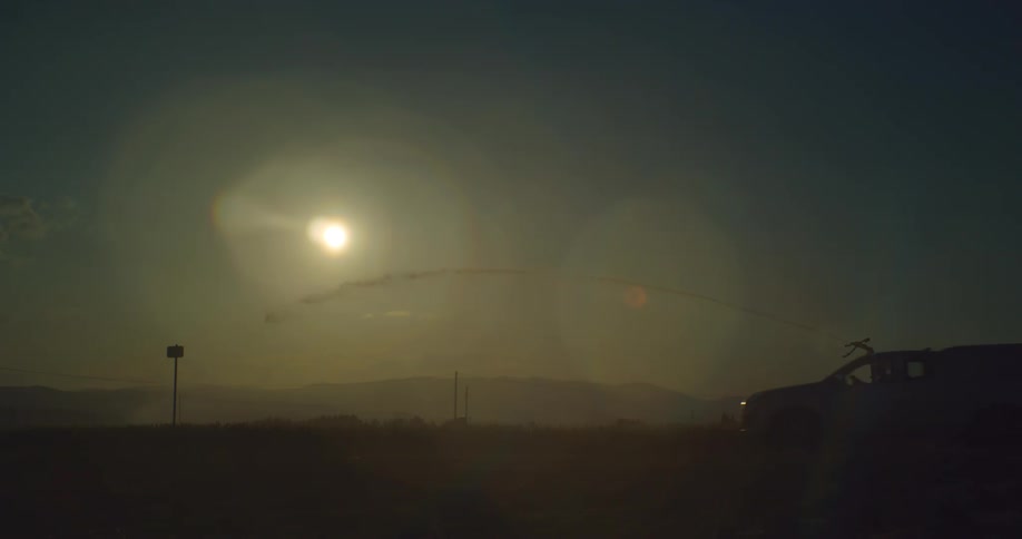 Sprinkler sprays a field at dusk as a truck passes by.