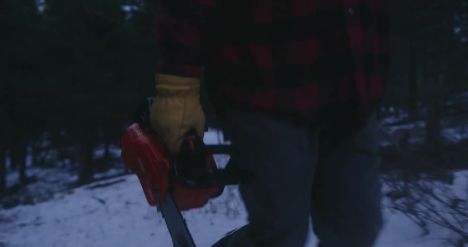 A man walks up a snowy hill carrying a chainsaw