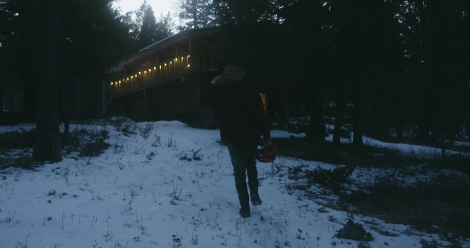 Man walks toward large cabin sitting in snow covered woods carrying a chainsaw and a log