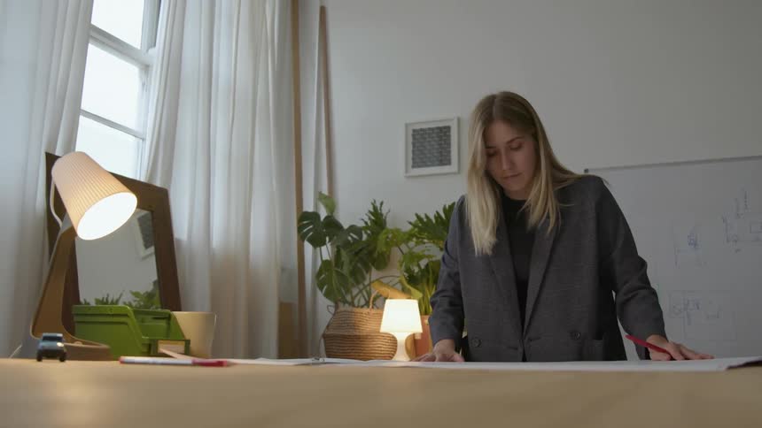 A woman straightens and studies a large set of plans with a white board hanging behind her