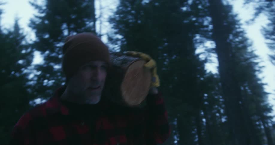 A man in a flannel shirt carrying a log through a forested area at blue hour