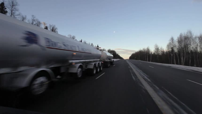 A semi-truck drives on a tree-lined road at sunset