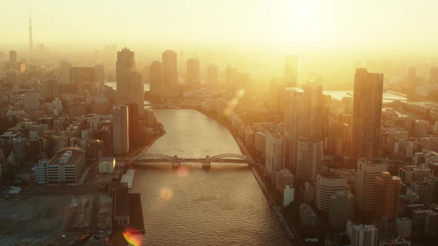 Traffic moves across a bridge crossing a river outside a big city with tall buildings and skyscrapers as the sun shines brightly in the hazy sky