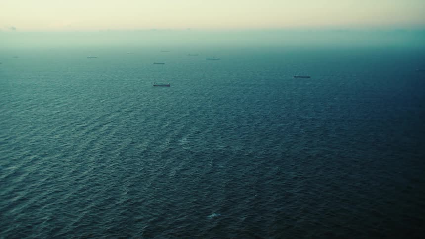 The sun shines on ripples and waves in the water as ships sail under a hazy sky