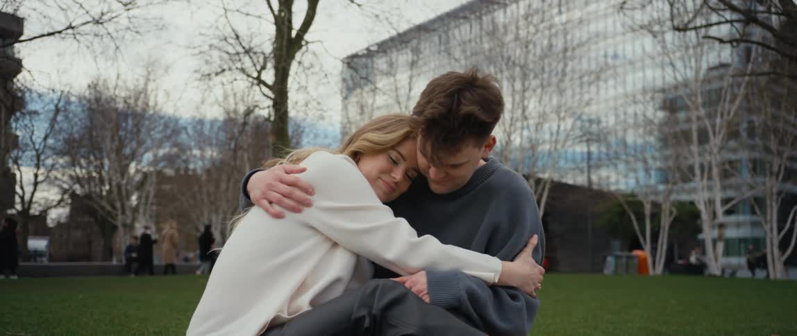 A woman rubs a mans chest as they hug in a public park as people walk behind them