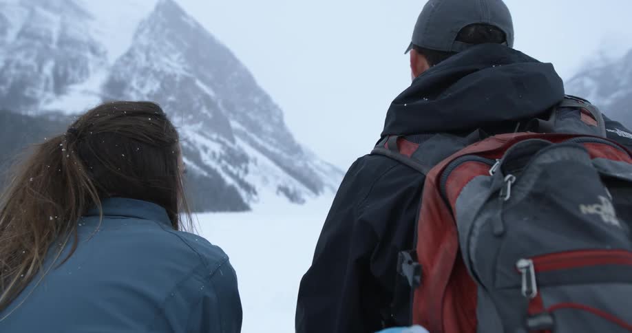 A man and woman are standing before a mountain as it snows