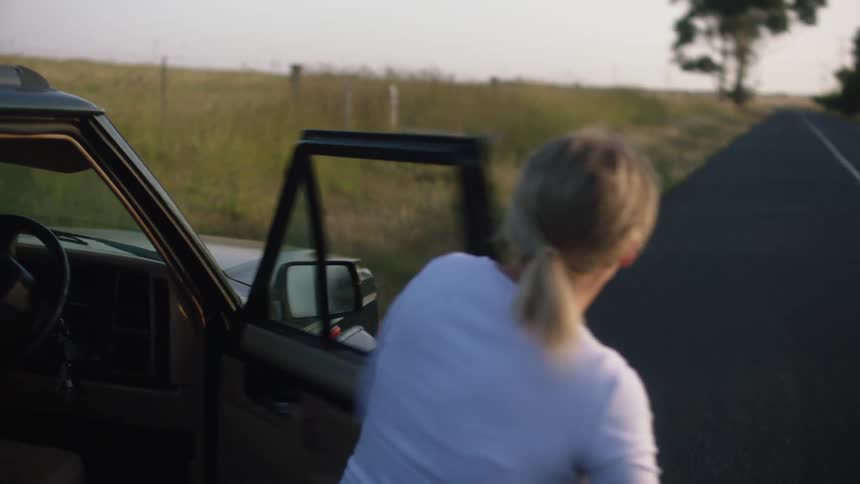 A woman jumps out of a vehicle parked on the side of a rural road and runs to the front and opens the hood