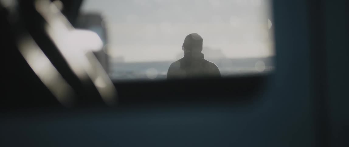 Person wearing glasses and a toque and coat standing outside a window looking at a lake