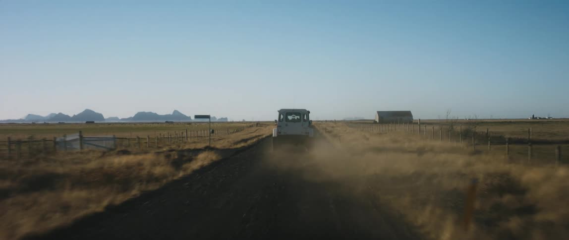 A vehicle drives down a dirt road through flat fields