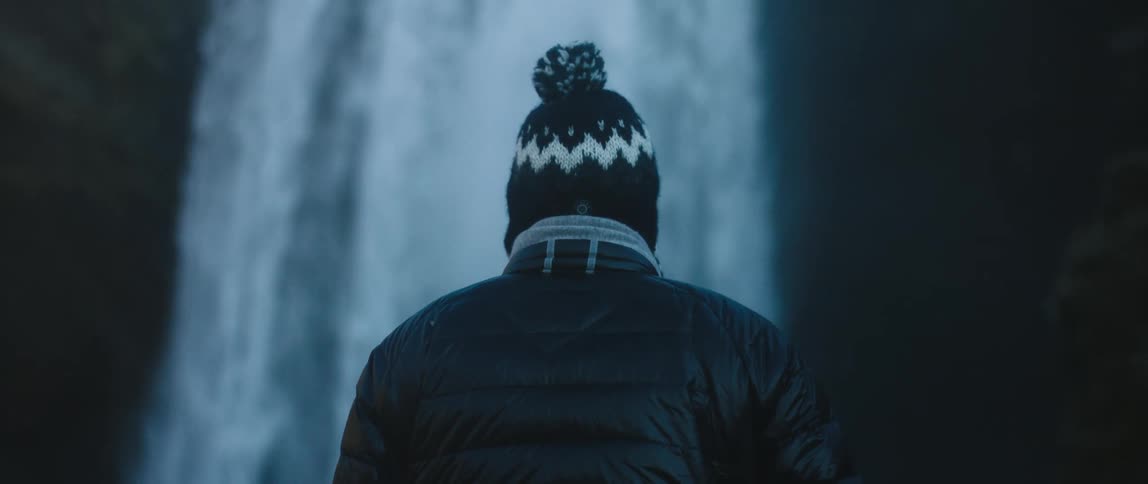 Person wearing knit hat and jacket stands looking up at waterfall flowing down cliff