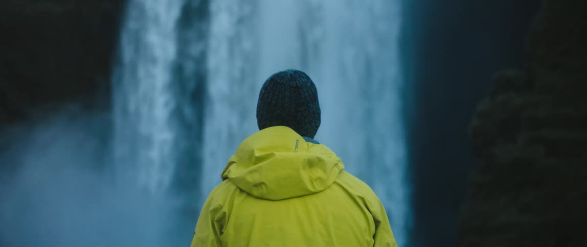 Man with woolly hat and yellow raincoat watches waterfall