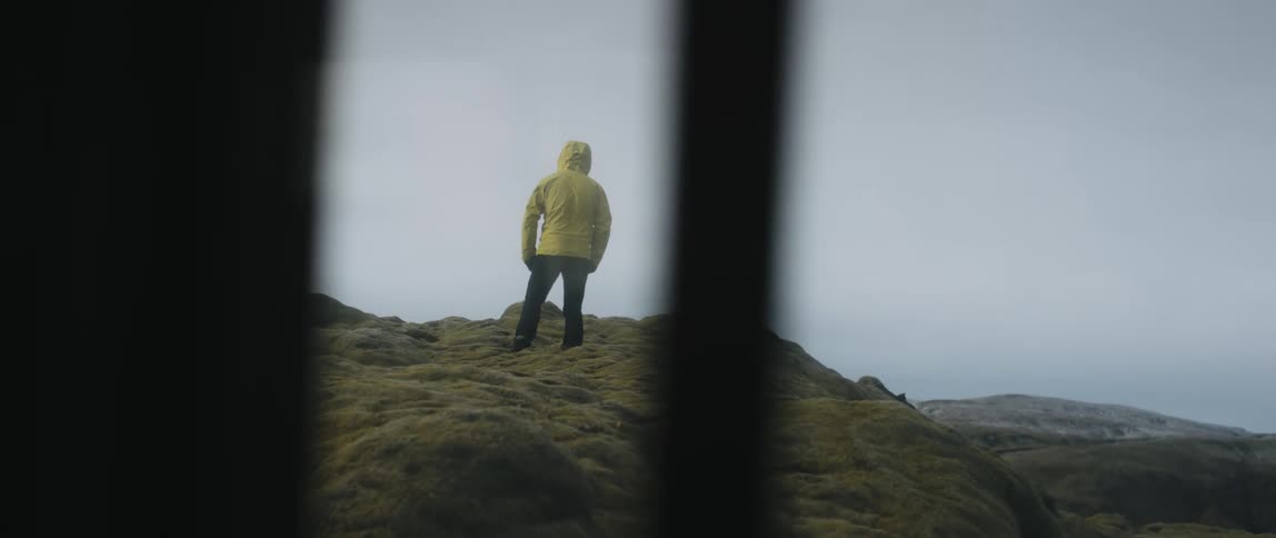 A man wearing a yellow rain jacket braces himself against the wind while standing on a rocky ridge