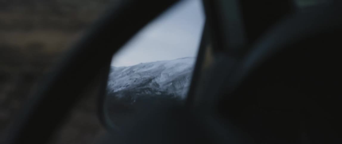 Person drives car through countryside as side mirror reflects snow covered mountain