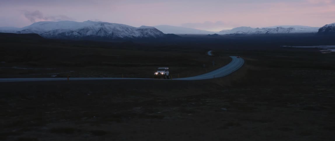 White vehicle travels on the highway with fields on each side and snowy mountains in the distance