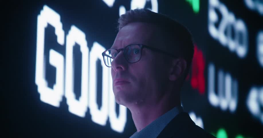 Clean shaven serious businessman looks at large screens with numbers