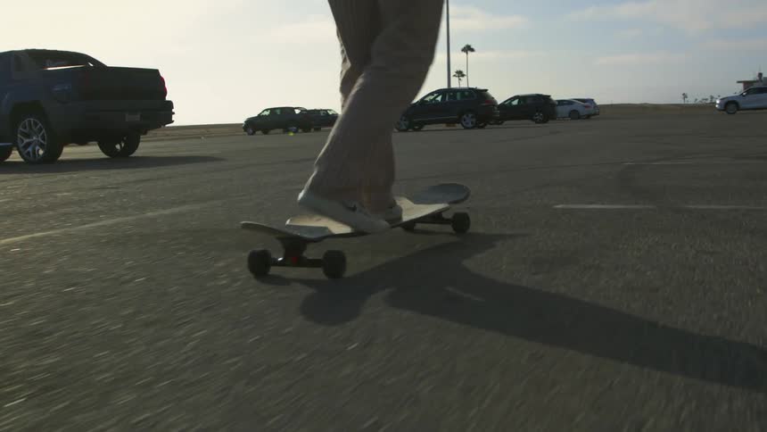 A person dances atop a skateboard while skating through a parking lot during the day