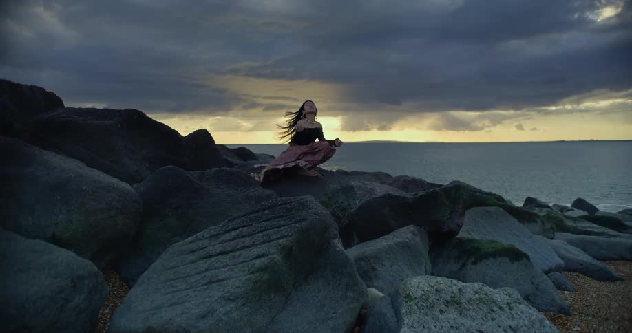 A woman kneels on rocks overlooking the ocean