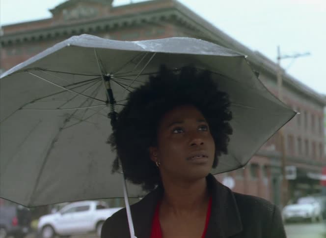 Woman with curly hair and gray coat carries umbrella walking down the street noticing something in the city