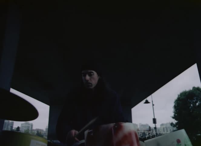 Male drummer with long hair and black hat and sweater plays drums under the bridge