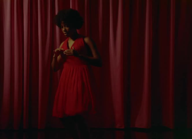 Woman with curly hair and red dress dances in front of red curtains on the stage
