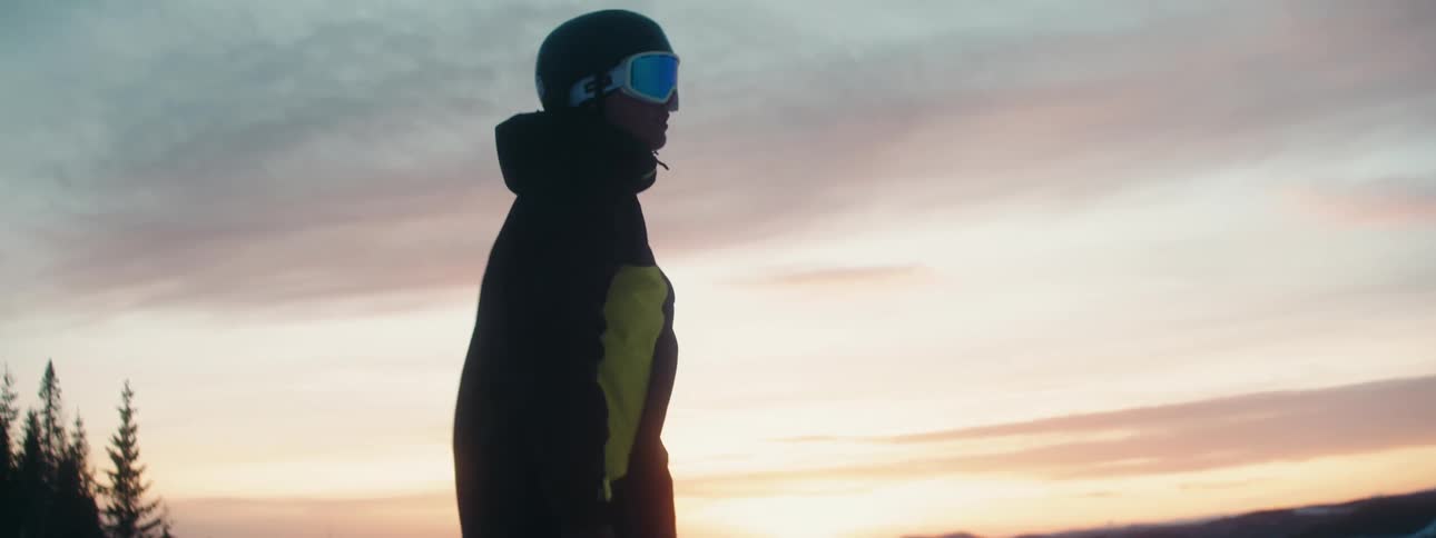 Person wearing goggles and a helmet stands on top of snowy slope as sun sits on horizon in red sky