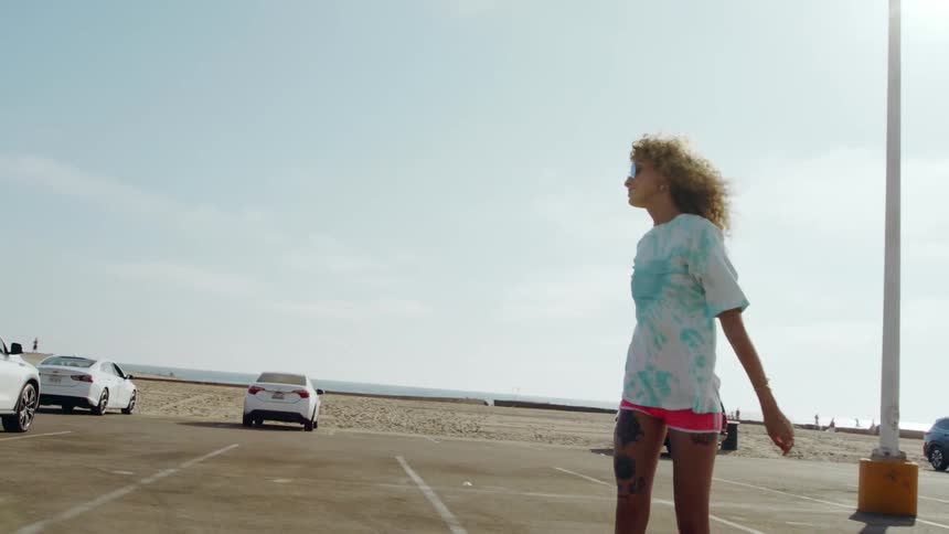 Female skateboarder with curly hair pink shorts and sunglasses skateboards in car park next to beach