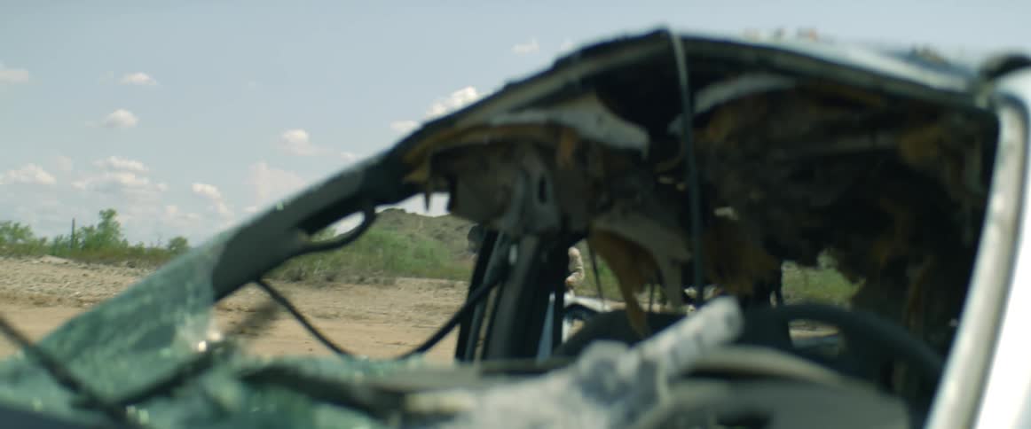 Soldiers approach a burnt down vehicle in a gravel lot