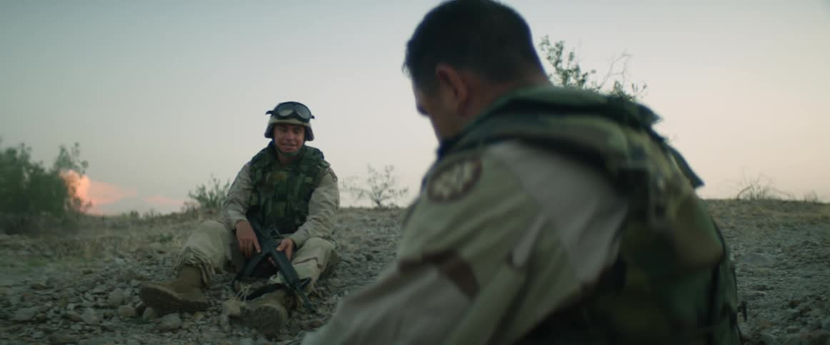 Two military soldiers are sitting along a rocky hillside taking a break and talking to each other