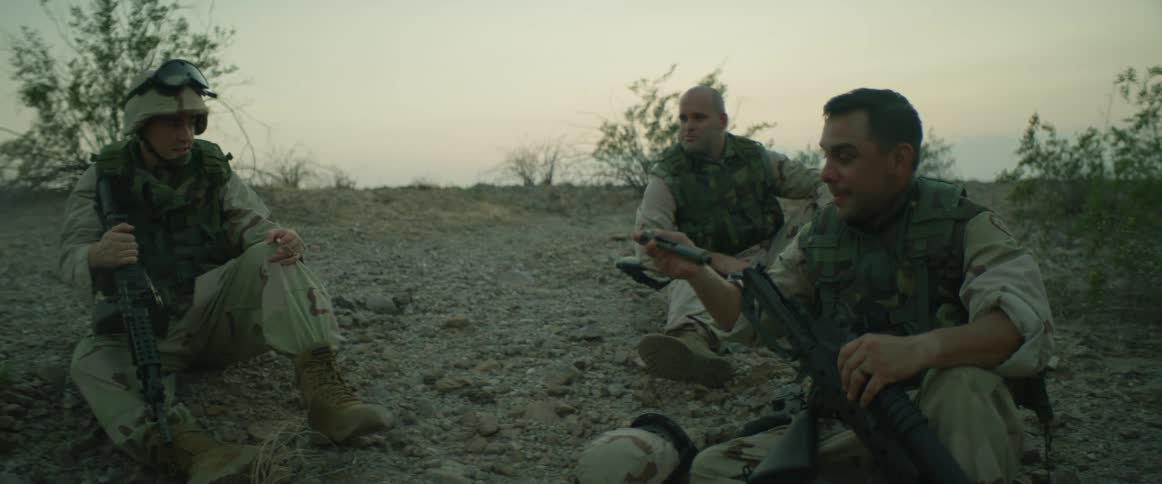 Three military soldiers are sitting along a rocky hillside taking a break and one of the men is taking apart his machine gun and hands parts to the man behind him