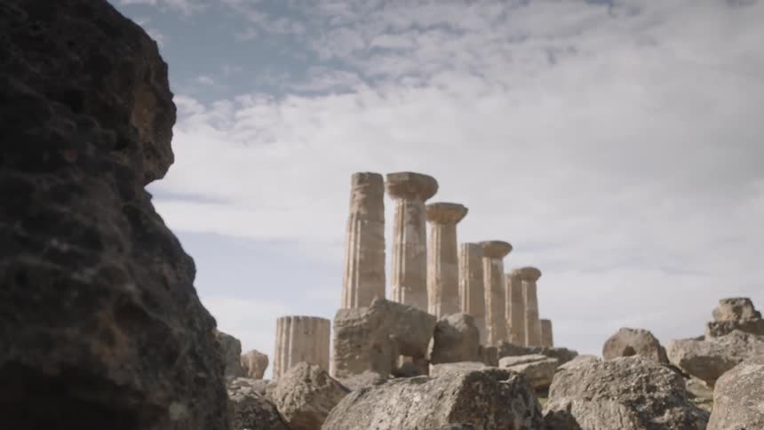 Greek ruins with tall columns stand in the distance against a cloudy sky