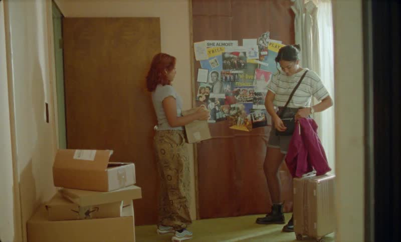 Friends moving out of a house stand in front of a board filled with pictures and flyers inside a room