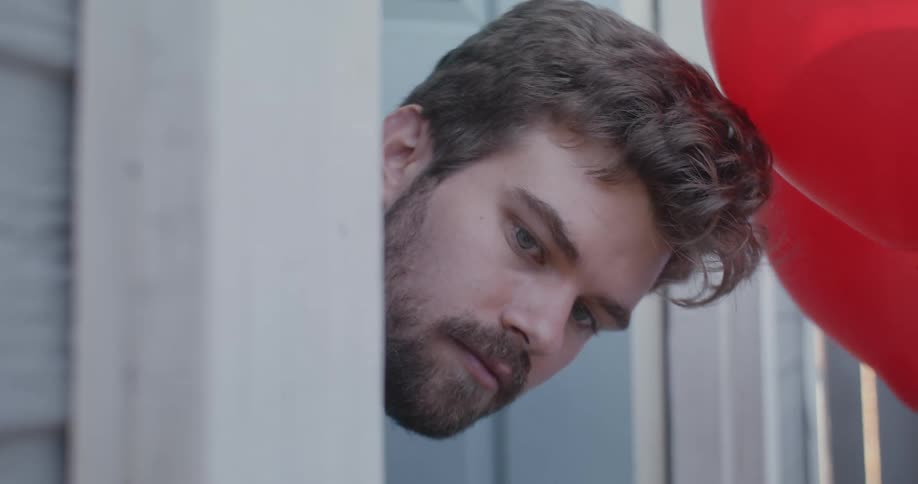 A man sticks his head out of the door of his house and sees red balloons floating in front of his face