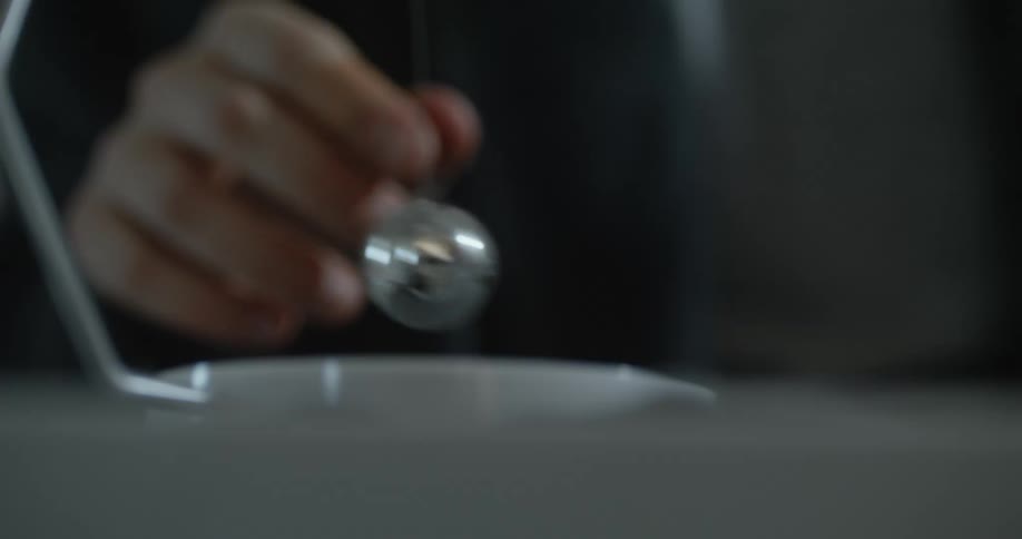 Light shines on the silver sphere of a pendulum as someone places it on a table and pushes the ball
