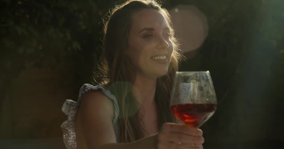A woman sitting outside in the sunshine clinks her wine glass with another in a toast