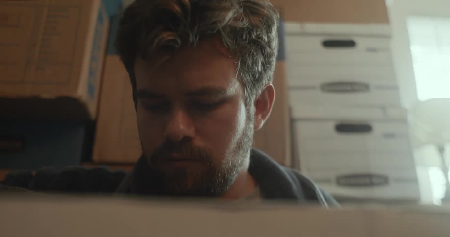 A white man is sitting down indoors and looking at stuff inside of a box