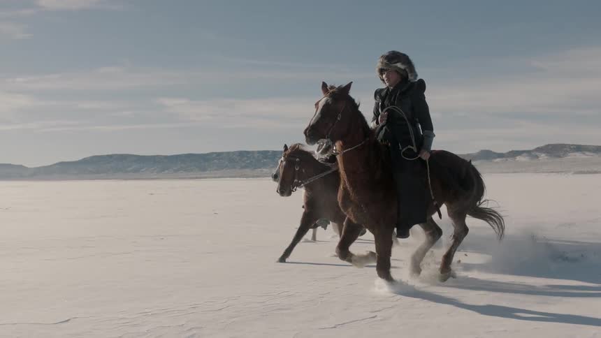 Three riders in mongolian-style clothing run their horses across a snowy plain