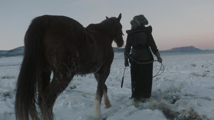 A man in heavy clothing walks a horse through the snow at dusk