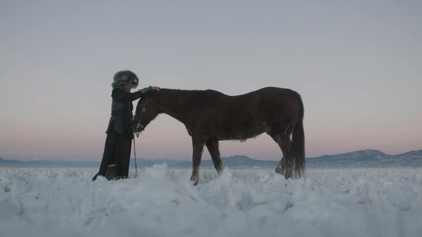 A man in heavy clothing pets his horse while standing in the snow