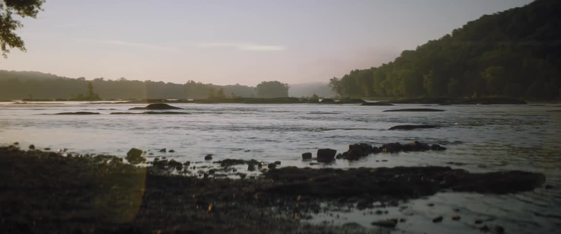 Water ripples as it flows along the shore with a forest of trees along the coast under a hazy sky