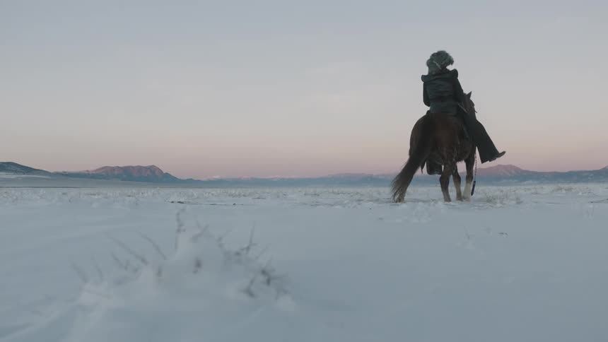 A man in heavy clothing mounts a horse in the snow