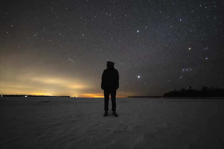 A silhouette of a person standing in the snow watching a sky filled with stars