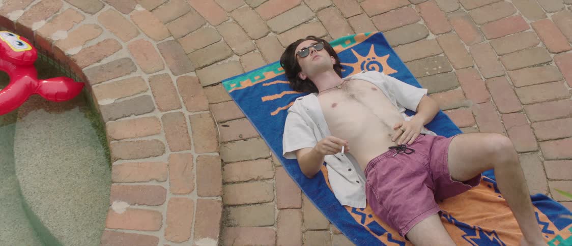 A man smoking a cigarette while lying on a beach towel next to a swimming pool