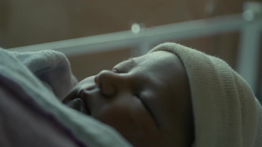 A newborn is wrapped in a blanket and has a hat on as it sleeps in a crib